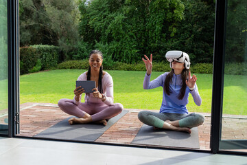 Diverse female friends sharing tablet on yoga mats on patio while wearing VR headset and headphones