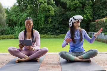 Diverse female friends practicing wellness on yoga mats on brick patio using tablet and VR headset