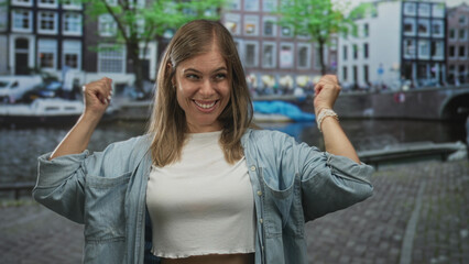 Fototapeta premium Young blonde woman raises clenched fists in front of canal water and brick buildings on street in amsterdam; victory joy.