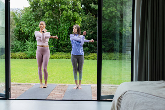Diverse female workout partners stretching across chest on back patio with yoga mats and smartwatch - Powered by Adobe