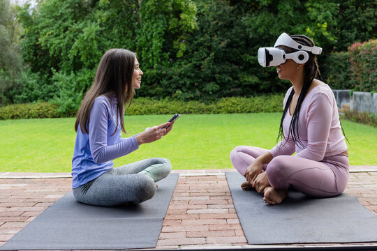 Diverse female friends sitting on yoga mats on backyard patio with smartphone and VR headset - Powered by Adobe