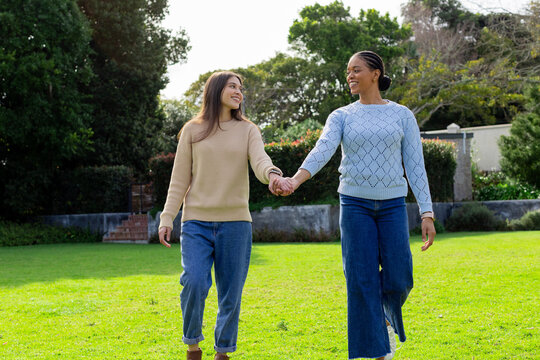 Diverse women walking hand in hand across garden lawn beside stone wall shrubs and brick steps