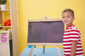 Boy child standing right beside easel chalkboard on blue table facing camera chalk, copy space