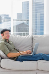 Mid-adult man reclining on left of light-gray sofa in modern living room using laptop, copy space