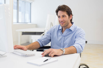 Mid-adult man sitting in wheelchair at white desk in bright office using monitor and keyboard
