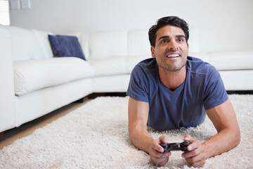 Man lying on shag rug holding wired game controller wearing blue tee in living room