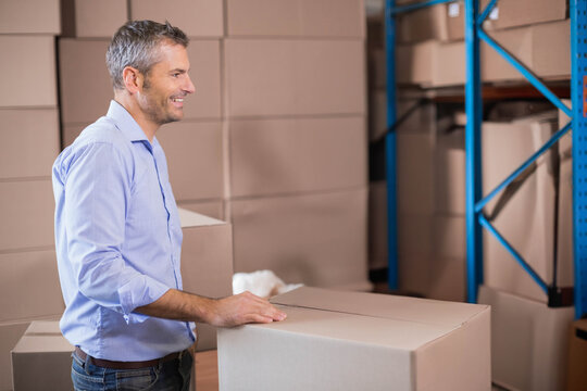 Middle-aged man in blue shirt and jeans stacking sealed boxes on blue metal shelving in warehouse