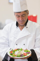 Male chef wearing tall white hat and white jacket presenting shrimp salad plate looking in kitchen