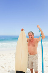 Naklejka premium Senior man shirtless standing on sandy beach holding off-white and blue surfboards by calm ocean