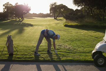 Mature man bending, placing golf ball on tee at sunlit course with white cap glove bag