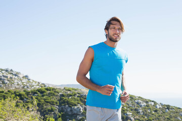 Man in mid twenties running along rocky coastal hillside trail wearing bright blue sleeveless top