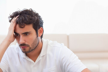 Obraz premium Man sitting on beige sofa at home wearing white polo, holding head and looking down