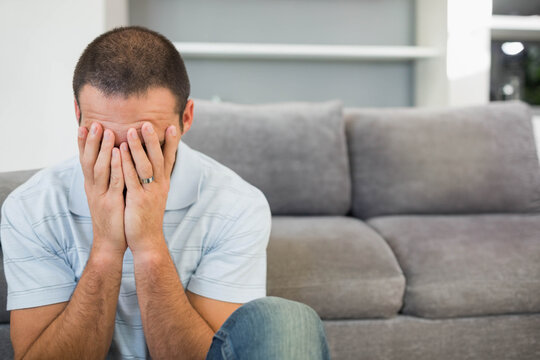 Mid-adult man sitting on gray sofa in living room covering face, wedding band visible, copy space