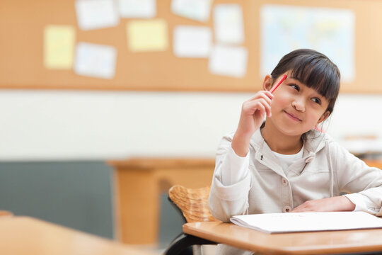 Asian child sitting in classroom at desk holding red pencil, open notebook with cork board visible