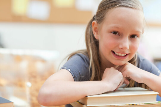 Female child student leaning on stack of hardcover books at classroom desk, wearing grey tee