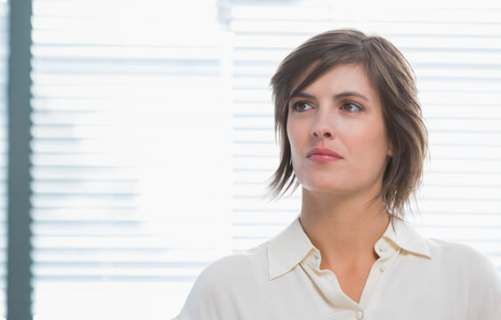 Woman mid 30s standing by backlit window with blinds in office wearing cream blouse, copy space