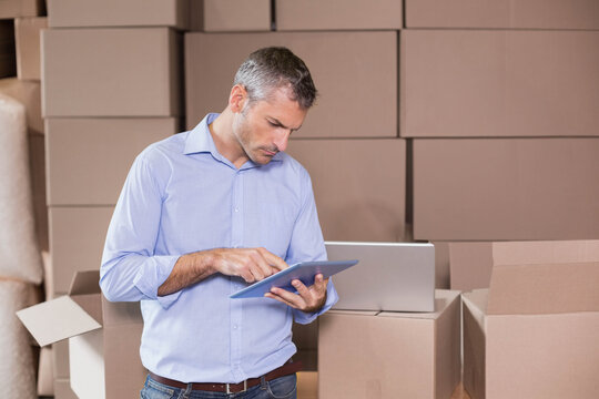 Mid-adult man wearing blue shirt, jeans working on tablet among boxes with laptop in warehouse - Powered by Adobe