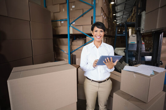 Female warehouse supervisor standing and tapping tablet in aisle with boxes papers in white shirt