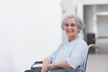 Senior female patient sitting in wheelchair smiling while wearing blue gown in clinic, copy space