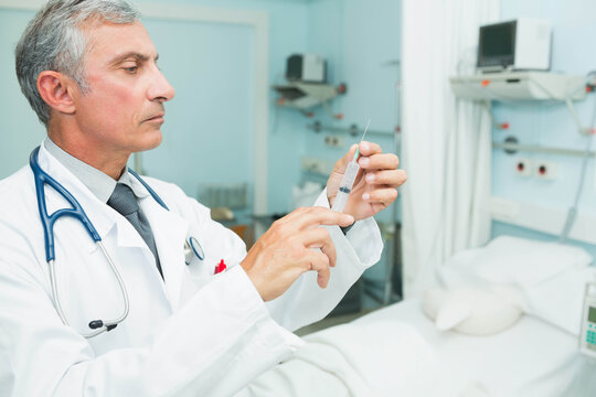 Senior male physician standing in lab coat at bedside preparing syringe with vial, stethoscope