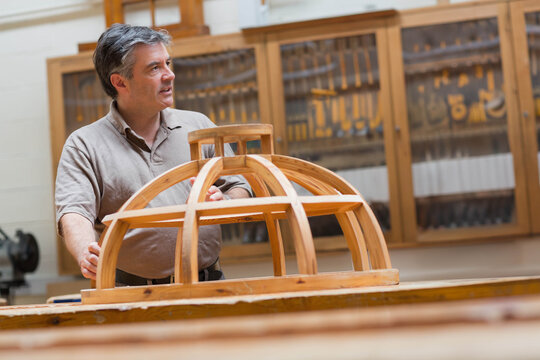 Mature man standing behind wooden dome framework at workshop, inspecting workbench tools