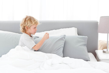 Child boy sitting on bed holding small object in right hand amid white duvet, copy space