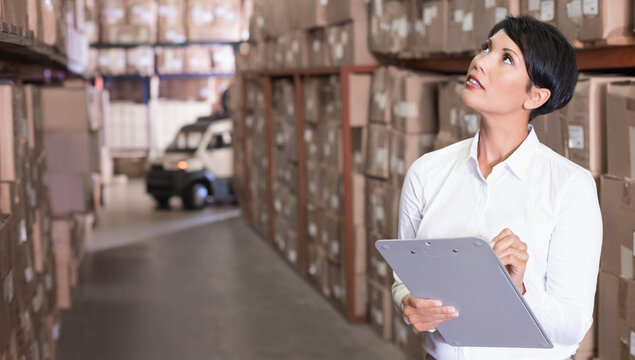 Mature woman in white shirt holding clipboard, pen checking boxes in storage aisle, copy space