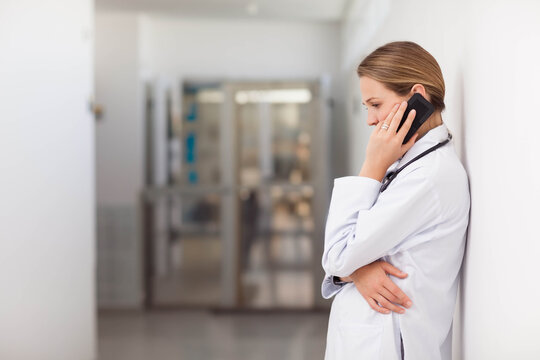 Female doctor wearing lab coat, leaning on right hospital corridor, holding smartphone, copy space