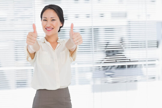 Asian woman giving double thumbs-up before venetian blinds in office wearing cream blouse and skirt