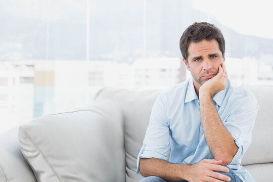 Man wearing blue shirt sitting on sofa, pressing ringed hand to face, gazing windows, copy space