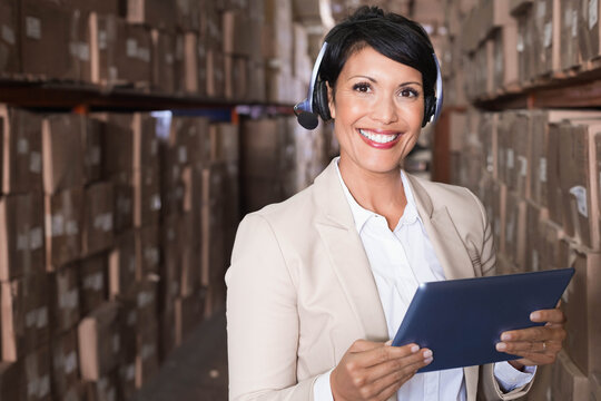 Mature female manager standing in narrow warehouse aisle holding dark tablet and using headset - Powered by Adobe