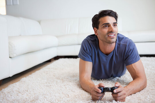 Mid-adult man lying on white shag rug in living room wearing blue t-shirt, holding wired controller