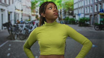Woman with exposed midriff standing hands on hips and pointing finger up on a city street during daytime; urban curiosity.