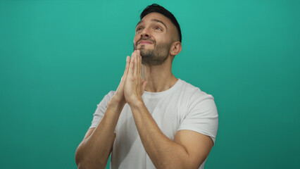 Young man with beard in white shirt praying with hopeful expression against green background