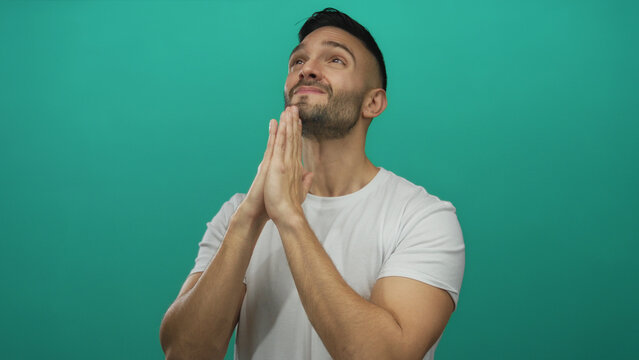 Young man with beard in white shirt praying with hopeful expression against green background