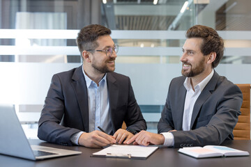 Two smiling businessmen discussing and finalizing a legal document during a corporate meeting in a modern office, symbolizing partnership, agreement, and successful deal making