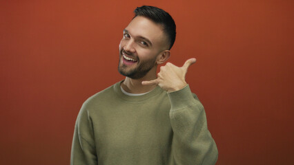 Fototapeta premium Young hispanic man in casual sweater gestures call me with a friendly smile against a vibrant orange background, exhibiting positive communication and modern style.