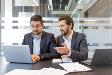 Two businessmen discussing project details and analyzing data on a laptop, fostering teamwork and professional collaboration in a modern office environment