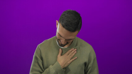 Young man with a beard in a green sweater showing gratitude against a vibrant purple background, expressing a heartfelt gesture by placing a hand on his chest.