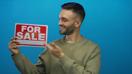 Young man smiling holding a red for sale sign against a bright blue background, suggesting real...