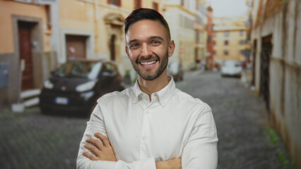 Fototapeta premium Smiling hispanic man standing confidently with crossed arms in an urban street of an old town city wearing a white shirt outdoors showcasing an urban background.