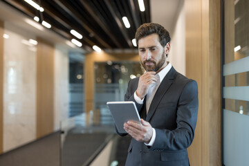 Confident businessman standing in a contemporary office corridor, holding a digital tablet while thinking and strategizing for business success and technological integration