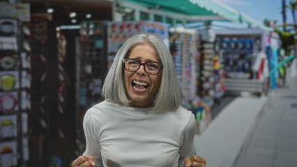 Middle aged woman with clenched fists shouting in a busy street souvenir market aisle, grey hair...