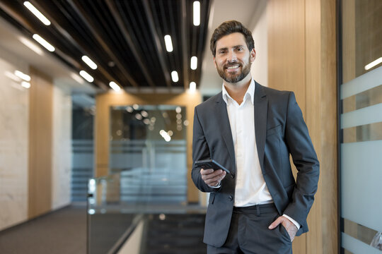Confident businessman in a modern office corridor holding a smartphone, smiling at camera, conveying success, connectivity and professional communication in a corporate environment