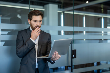 Fototapeta premium Worried businessman holding a laptop, expressing shock and concern while standing in a modern office environment with glass partitions, reflecting a problem or challenge