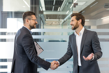 Two smiling business professionals in formal wear are shaking hands in a modern office environment, symbolizing a successful deal, partnership, trust, and collaboration