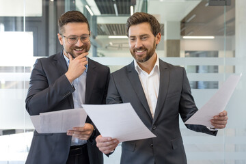 Two bearded businessmen in suits smiling and reviewing financial reports together in a modern office, collaborating on strategy, planning and paperwork during a meeting