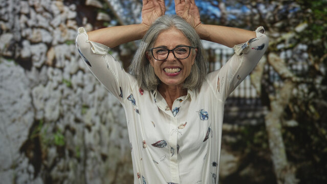 Woman holding hands on head smiling with glasses and white feathered blouse in forest near stone gate; playful mischief.
