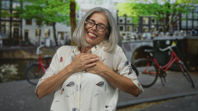 Middle aged grey haired woman with hands on chest and open arms on street in amsterdam; gratitude warmth.