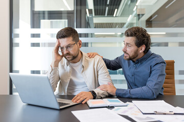 Two male coworkers in a modern office, one offering comfort and support to the other who is...